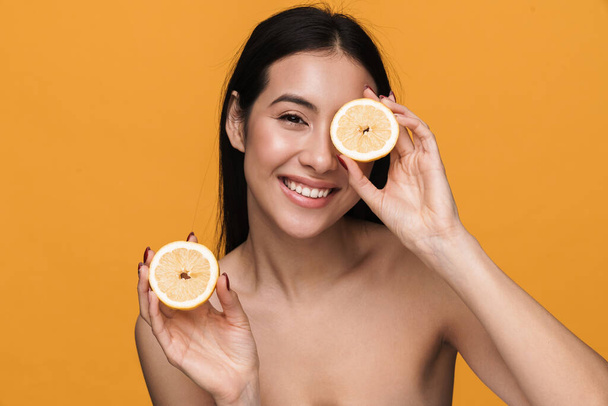 Beauty portrait of caucasian young brunette half-naked woman smiling and holding lemon parts isolated over yellow background - 写真・画像