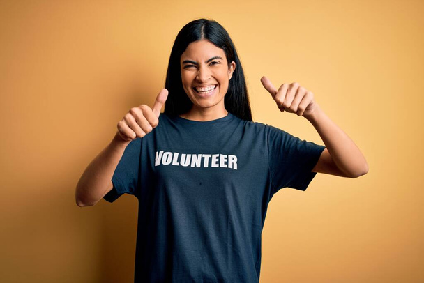 ボランティアのTシャツを身に着けている若い美しいヒスパニック系の女性は、社会的慈善道徳的な承認手で積極的なジェスチャーを行う,親指を笑顔と成功のために幸せ.勝者のジェスチャー. - 写真・画像