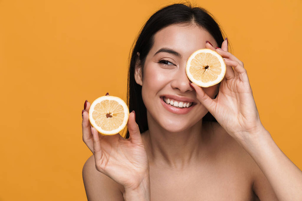 Beauty portrait of caucasian young brunette half-naked woman smiling and holding lemon parts isolated over yellow background - Foto, Bild