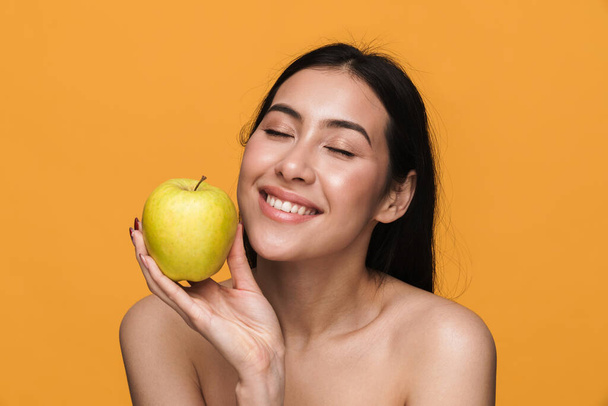 Beauty portrait of caucasian young brunette half-naked woman smiling and holding green apple isolated over yellow background - Photo, Image