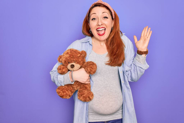 Jonge roodharige zwangere vrouw verwacht baby houden teddy beer over paarse achtergrond erg blij en opgewonden, winnaar uitdrukking vieren overwinning schreeuwen met een grote glimlach en opgeheven handen - Foto, afbeelding