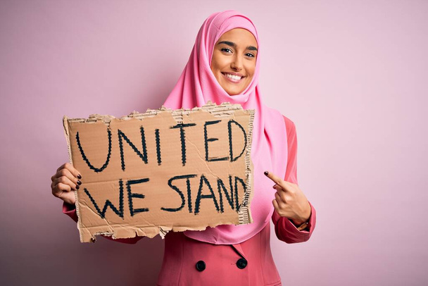 Giovane attivista donna indossa hijab rosa musulmano tenendo banner con uniti siamo messaggio molto felice che punta con mano e dito
 - Foto, immagini