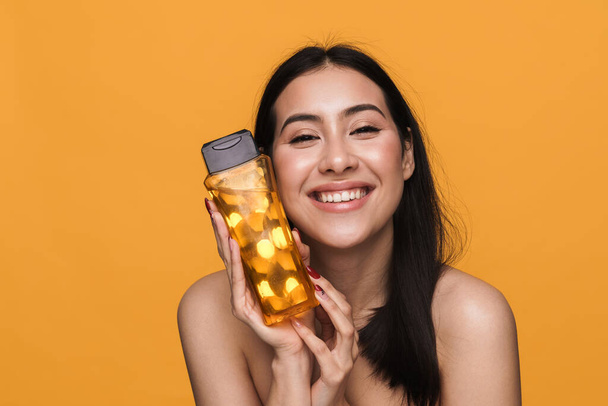 Beauty portrait of caucasian young brunette half-naked woman smiling and holding shower gel isolated over yellow background - Фото, зображення