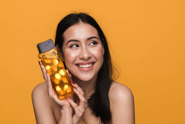 Beauty portrait of caucasian young brunette half-naked woman smiling and holding shower gel isolated over yellow background - Φωτογραφία, εικόνα