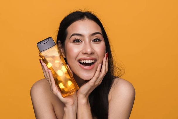 Beauty portrait of caucasian young brunette half-naked woman smiling and holding shower gel isolated over yellow background - Fotó, kép