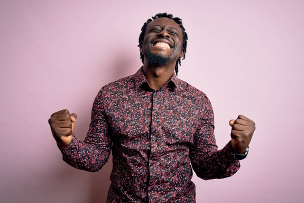Mladý pohledný africký Američan v neformální košili stojící nad růžovým pozadím velmi šťastný a vzrušený dělá vítězný gesto se zdviženýma rukama, usmívá se a křičí po úspěchu. Koncept oslavy. - Fotografie, Obrázek