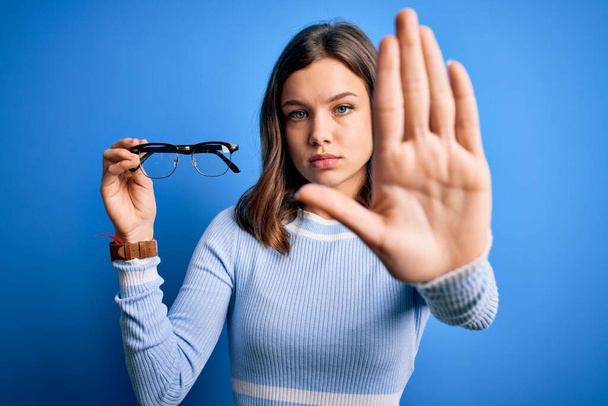 Jeune fille blonde tenant des lunettes lentille optique sur fond bleu isolé avec la main ouverte faisant stop signe avec expression sérieuse et confiante, geste de défense
 - Photo, image