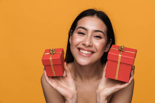 Beauty portrait of caucasian young brunette half-naked woman smiling while holding present box isolated over yellow background - Photo, Image