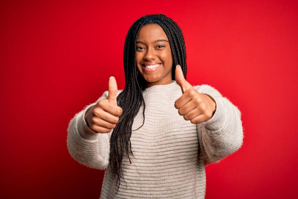 赤い隔離された背景の上にカジュアルな冬のセーターを着ている若いアフリカ系アメリカ人女性は、手で積極的なジェスチャーを行うことを承認し、親指を上げて笑顔と成功のために幸せ。勝者のジェスチャー. - 写真・画像