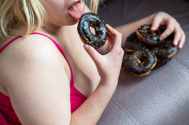 Fat blonde eats a chocolate donut. Unrecognizable obese young woman enjoying sweet buns for breakfast. Cropped photo. - Foto, Imagem
