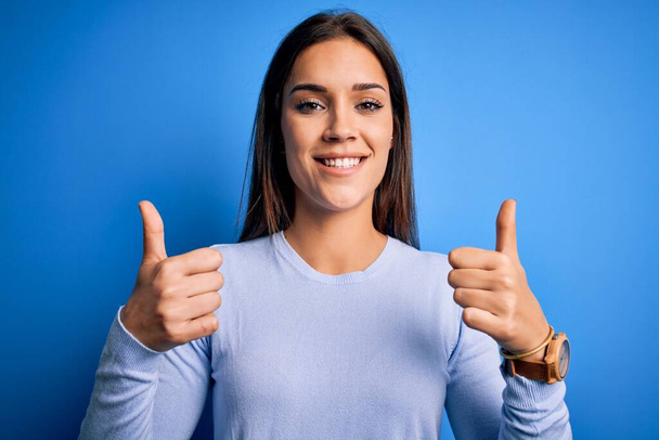 青の背景の成功の印の上に立つカジュアルセーターを身に着けている若い美しいブルネットの女性は手で積極的なジェスチャーをして、親指を笑顔と幸せ。陽気な表情と勝者のジェスチャー. - 写真・画像
