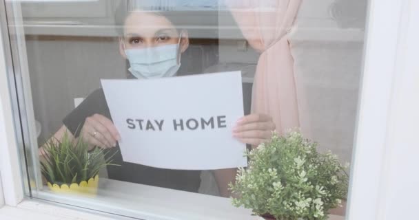 Young girl in medical mask, being at home, sitting near the window, and looking outside through the glass. Woman near window quarantined during pandemic covid-19, shows an inscription stay at home - 映像、動画