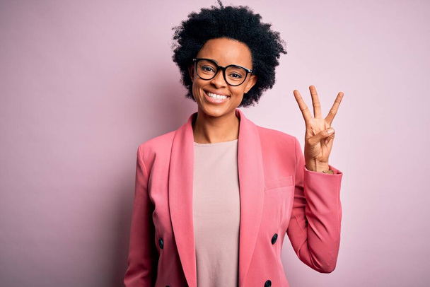 Молодая красивая афро-американка афро-бизнесмен с вьющимися волосами в розовой куртке показывает и указывает пальцами номер три, улыбаясь уверенно и счастливо
. - Фото, изображение