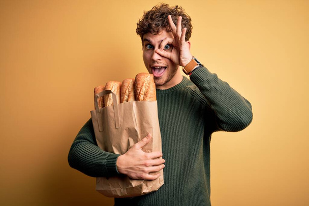 Young blond man with curly hair holding paper bag with bread over yellow background with happy face smiling doing ok sign with hand on eye looking through fingers - Photo, Image