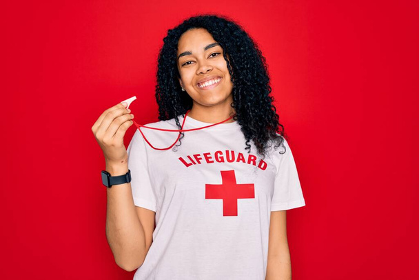 Jonge Afro-Amerikaanse gekrulde badmeester vrouw draagt t-shirt met rood kruis met behulp van fluitje met een vrolijk gezicht staan en glimlachen met een zelfverzekerde glimlach tonen tanden - Foto, afbeelding