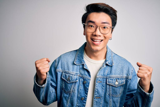 Jeune bel homme chinois portant une veste en denim et des lunettes sur fond blanc célébrant surpris et étonnés du succès avec les bras levés et les yeux ouverts. Concept gagnant
. - Photo, image