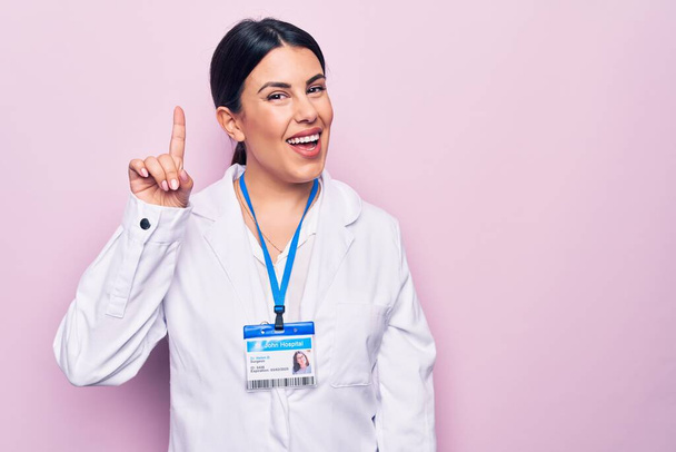 Νεαρή όμορφη γιατρός γυναίκα φορώντας στηθοσκόπιο και ταυτότητα πάνω από απομονωμένο ροζ φόντο χαμογελώντας με μια ιδέα ή ερώτηση δείχνοντας το δάχτυλο προς τα πάνω με χαρούμενο πρόσωπο, νούμερο ένα - Φωτογραφία, εικόνα