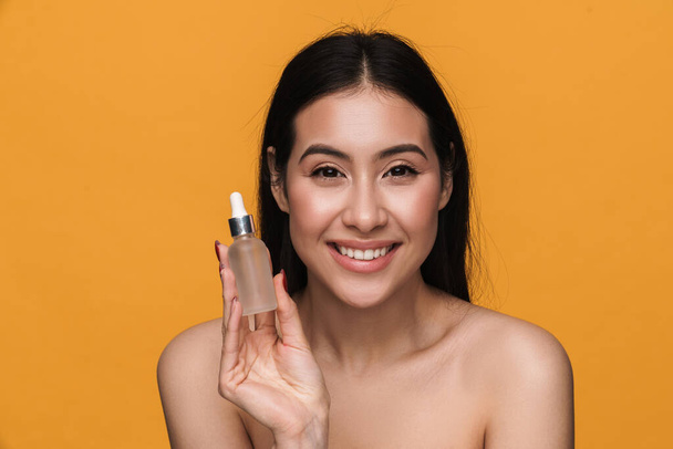 Beauty portrait of young caucasian half-naked woman smiling and holding facial serum cream bottle isolated over yellow background - Фото, изображение