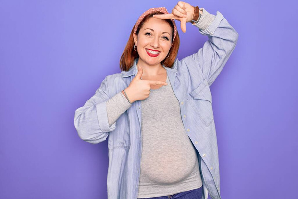 Jonge mooie roodharige zwangere vrouw verwacht baby over geïsoleerde paarse achtergrond glimlachen maken frame met handen en vingers met gelukkig gezicht. Creativiteit en fotografie concept. - Foto, afbeelding