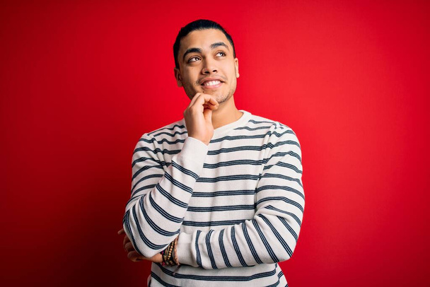 Young brazilian man wearing casual striped t-shirt standing over isolated red background with hand on chin thinking about question, pensive expression. Smiling and thoughtful face. Doubt concept. - Foto, Imagen