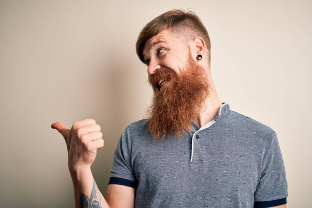 Нахабний ірландський рудий чоловік з бородою і татуюванням руки стоїть над ізольованим фоном посміхаючись з щасливим обличчям і вказуючи на бік з великим пальцем вгору. - Фото, зображення