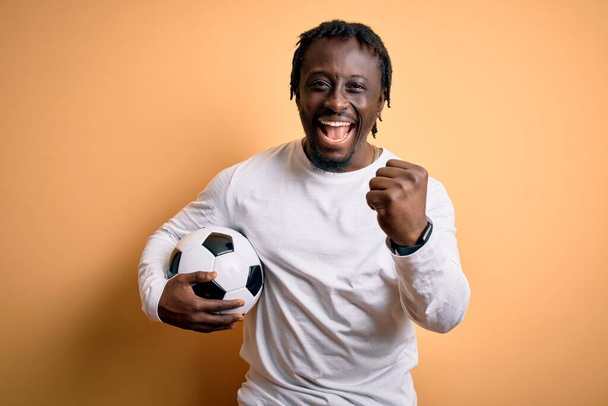 Giovane giocatore afro-americano uomo che gioca a calcio tenendo palla da calcio su sfondo giallo urlando orgoglioso e celebrando la vittoria e il successo molto eccitato, esultante emozione
 - Foto, immagini