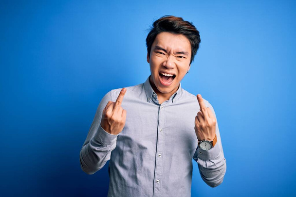 Joven hombre chino guapo con camisa casual de pie sobre fondo azul aislado Mostrando dedo medio haciendo mierda mala expresión, provocación y actitud grosera. Gritando emocionado
 - Foto, Imagen