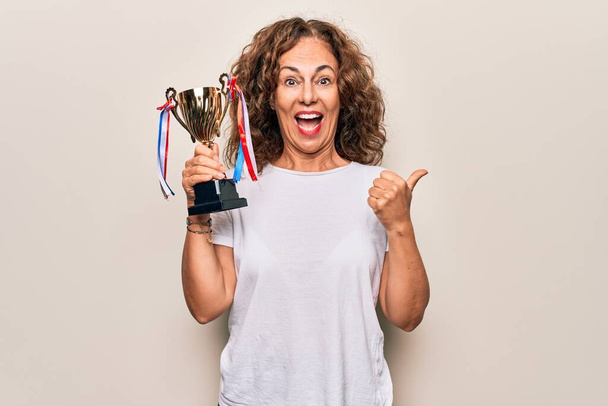 Middle age beautiful successful woman holding trophy for victory over white background pointing thumb up to the side smiling happy with open mouth - Foto, immagini