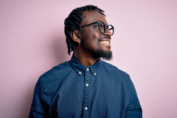 Mladý pohledný africký Američan v neformální košili a brýlích přes růžové pozadí, dívá se stranou s úsměvem na tváři, přirozený výraz. Smějící se sebevědomí. - Fotografie, Obrázek