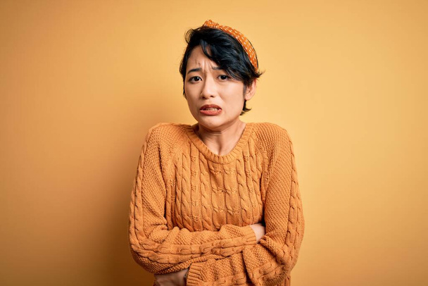Young beautiful asian girl wearing casual sweater and diadem standing over yellow background shaking and freezing for winter cold with sad and shock expression on face - Фото, изображение