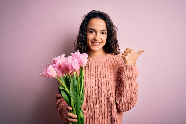 Młoda piękna romantyczna kobieta z kręconymi włosami trzymająca bukiet różowych tulipanów wskazując na plecy z ręką i kciukami w górze, uśmiechnięta pewna siebie - Zdjęcie, obraz