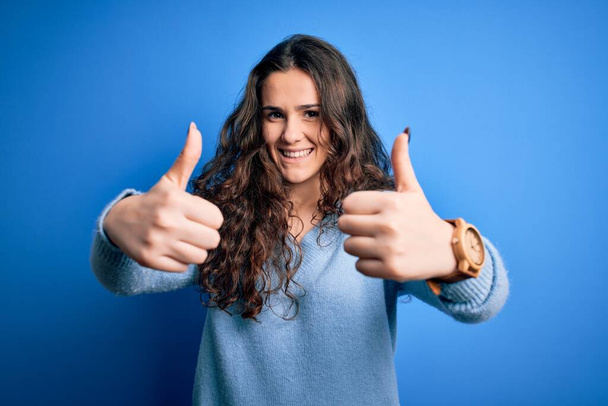 Jonge mooie vrouw met krullend haar dragen blauwe casual trui over geïsoleerde achtergrond goed te keuren doen positieve gebaar met de hand, duimen omhoog glimlachend en blij voor succes. Winnaar gebaar. - Foto, afbeelding