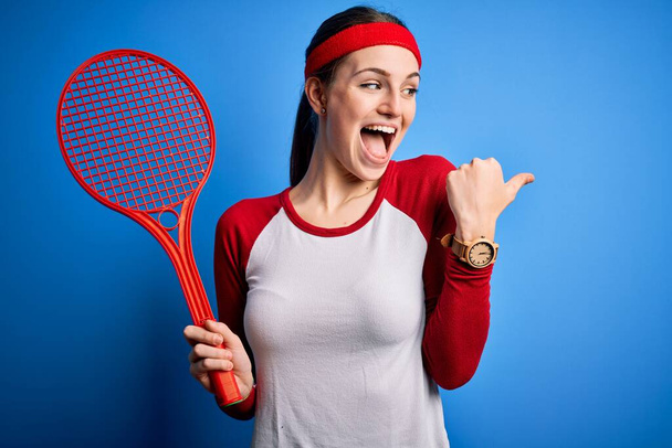 Νεαρή όμορφη κοκκινομάλλα αθλήτρια που παίζει τένις χρησιμοποιώντας ρακέτα πάνω από το μπλε φόντο δείχνοντας και δείχνοντας με τον αντίχειρα μέχρι το πλάι με χαρούμενο πρόσωπο χαμογελώντας - Φωτογραφία, εικόνα