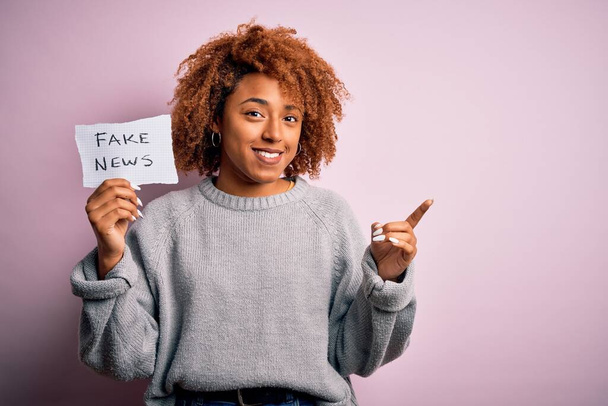 Mladá afro Afroameričanka s kudrnatými vlasy drží papír s falešnou zprávou velmi šťastně ukazuje rukou a prstem na stranu - Fotografie, Obrázek