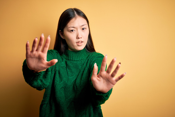 Sarı arka plan üzerine yeşil kazak giyen genç, güzel Asyalı kadın avuç içi hareketlerini durduruyor, kızgın ve hayal kırıklığına uğramış bir ifade takınıyor. - Fotoğraf, Görsel