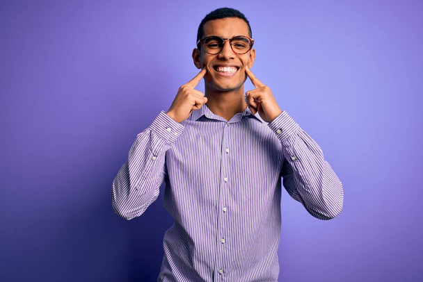 Schöner afrikanisch-amerikanischer Mann mit gestreiftem Hemd und Brille vor violettem Hintergrund, der mit offenem Mund lächelt, die Finger zeigt und ein fröhliches Lächeln erzwingt - Foto, Bild