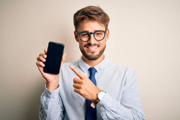 Young businessman holding smartphone standing over isolated white background very happy pointing with hand and finger - Photo, Image