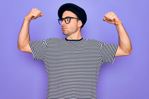 Bell'uomo con gli occhi azzurri che indossa t-shirt a righe e berretto francese su sfondo viola che mostra i muscoli delle braccia sorridenti orgogliosi. Concetto fitness
. - Foto, immagini