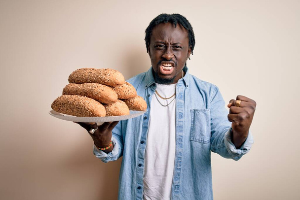 Fiatal afro-amerikai férfi kezében tálca egészséges teljes kiőrlésű kenyeret fehér háttér bosszús és frusztrált kiabált harag, őrült és kiabált emelt kézzel, harag koncepció - Fotó, kép