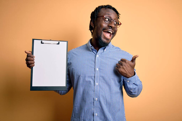 Молодой африканский американский инспектор в очках, держащий контрольный лист планшета, показывающий пальцем вверх в сторону, со счастливым лицом, улыбающимся
 - Фото, изображение