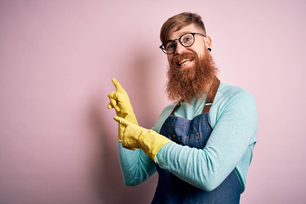 Ιρλανδός κοκκινομάλλης καθαριστής με γενειάδα που φοράει ποδιά και πλένει γάντια χαμογελώντας και κοιτάζοντας την κάμερα που δείχνει με τα δύο χέρια και τα δάχτυλα στο πλάι. - Φωτογραφία, εικόνα