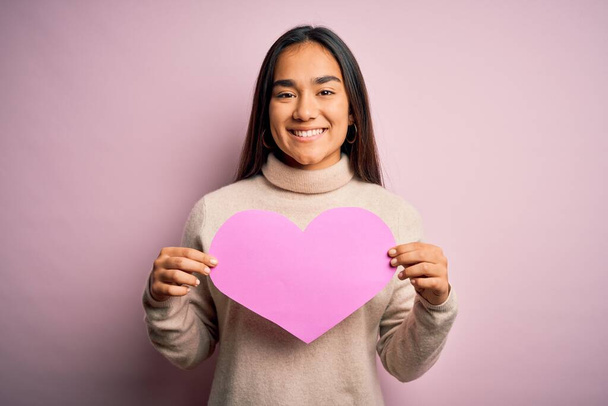 Young beautiful asian woman holding pink heart standing over isolated background with a happy face standing and smiling with a confident smile showing teeth - Zdjęcie, obraz