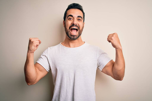 Young handsome man with beard wearing casual t-shirt standing over white background celebrating surprised and amazed for success with arms raised and open eyes. Winner concept. - Foto, Bild