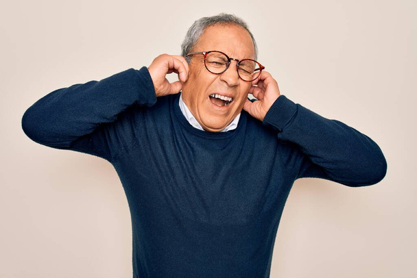 Старший красивий сіро-волохатий чоловік носить светр і окуляри над ізольованим білим тлом, покриваючи вуха пальцями з роздратованим виразом для шуму гучної музики. Концепція глухого
. - Фото, зображення