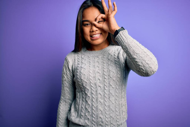 Jovem bela menina asiática vestindo camisola casual em pé sobre isolado roxo fundo fazendo ok gesto com a mão sorrindo, olho olhando através de dedos com rosto feliz
. - Foto, Imagem