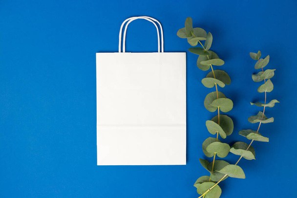 Biała papierowa torba z uchwytami i Eucalyptus Liście na niebieskim tle, płaskie ułożenie, widok z góry, przestrzeń do kopiowania, plastikowe przedmioty wolne - Zdjęcie, obraz
