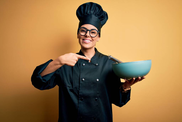 Giovane bella donna chef bruna indossa uniforme fornello e cappello tenendo ciotola e frusta molto felice puntando con mano e dito
 - Foto, immagini