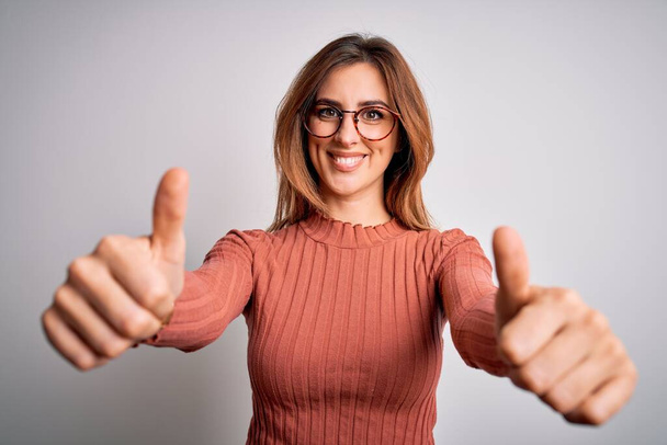 Young beautiful brunette woman wearing casual sweater and glasses over white background approving doing positive gesture with hand, thumbs up smiling and happy for success. Winner gesture. - Fotoğraf, Görsel