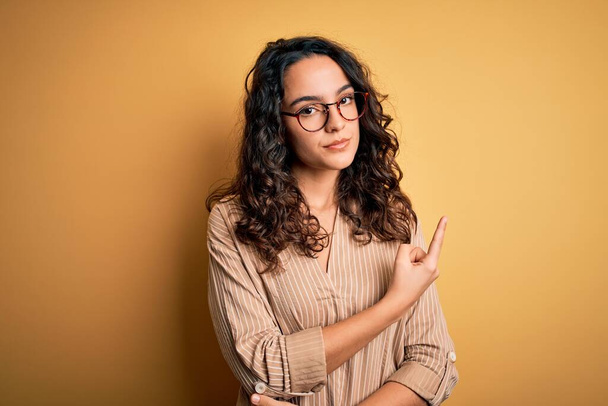 Beautiful woman with curly hair wearing striped shirt and glasses over yellow background Pointing with hand finger to the side showing advertisement, serious and calm face - Foto, Imagem