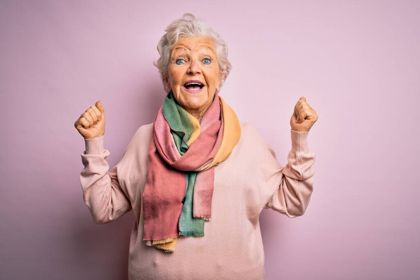 Senior bella donna dai capelli grigi indossa maglione casual e sciarpa su sfondo rosa celebrando sorpreso e stupito per il successo con le braccia alzate e occhi aperti. Concetto vincitore
. - Foto, immagini